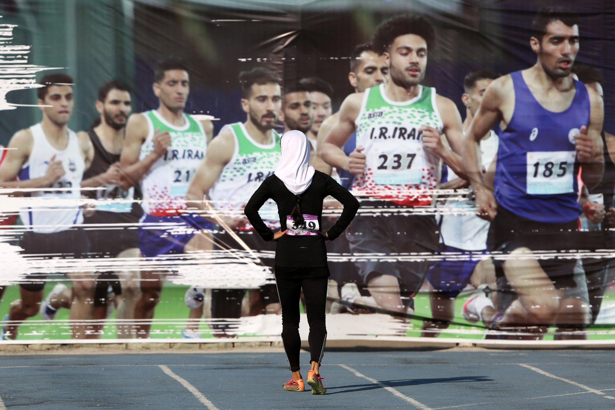 Picture shows a female athlete preparing to compete in the starting line with the mandatory hijab cover. She looks at pictures of male athletes in comfortable, standard competition clothes.