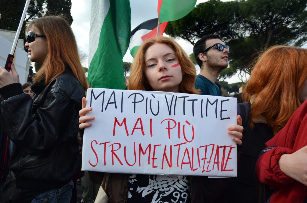 A woman protestor holding a sign "never again victims, never again exploited" in front of Palestinian flags. 