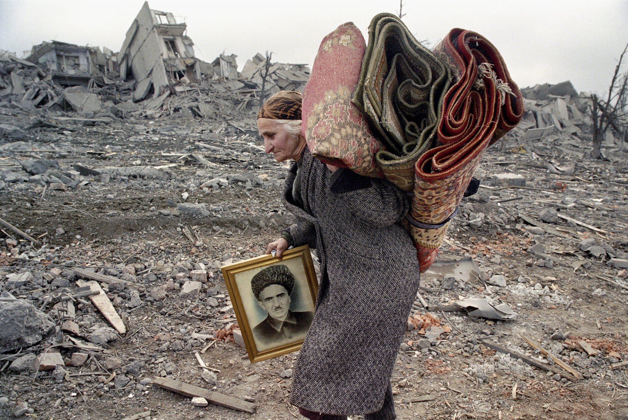 A Chechnyan woman carries a portrait of her husband and two carpets, while walking in the middle of ruins and rubble.