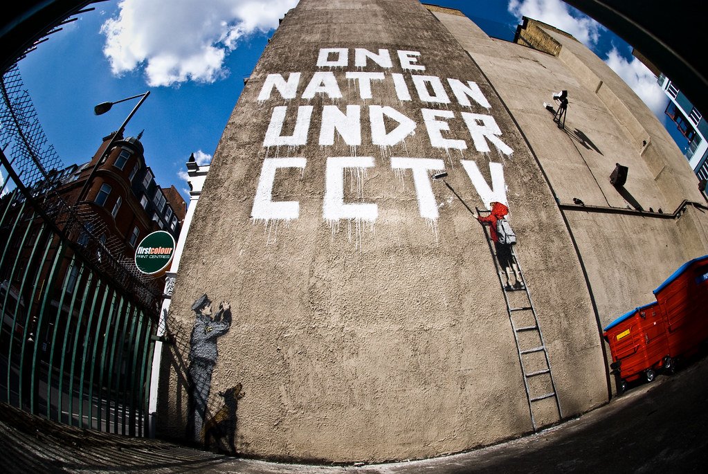 A mural depicting a child in a red hood painting “ONE NATION UNDER CCTV”, while being watched by a police officer and a dog. 