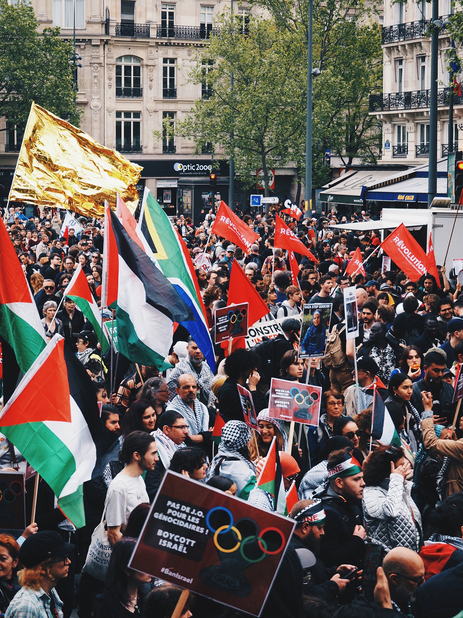 People hold Palestinian flags, "Revolution" flags, and banners against Olympics on Mayday demonstration in Paris.