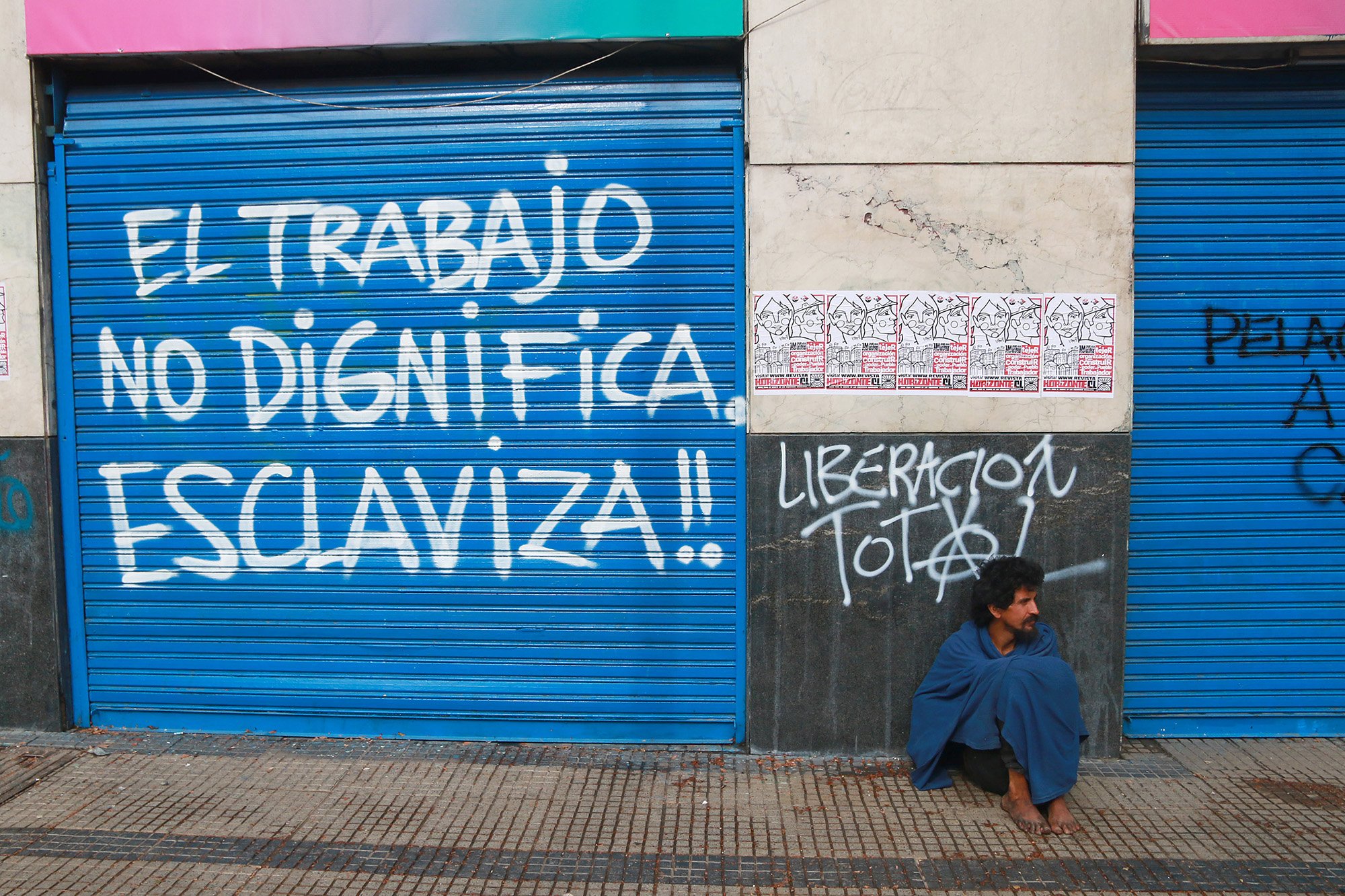 A homeless man sits by a wall with the slogan "Work does not dignify, it enslaves" written on it.     