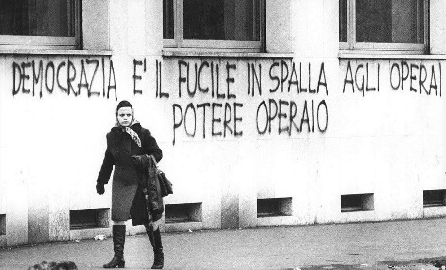 Italy, 1972: A woman passing by a wall on which a Potere Operaio slogan "Democracy is the rifle on the workers' shoulder" is written.