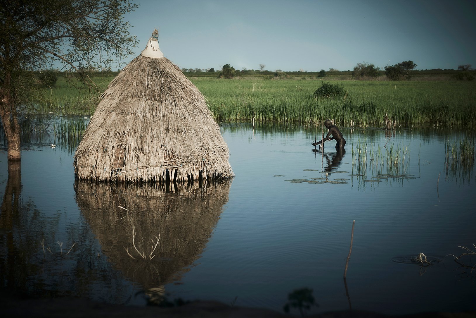 A man wades with a net to catch fish around Bentiu's floodwaters in front of flooded hut.