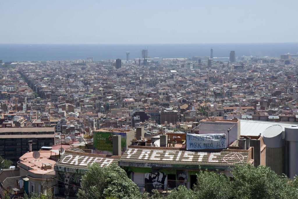 Visible from the top of Park Güell—a popular tourist destination—are prominent signs on a nearby apartment building reading "OKUPA Y RESISTE" (Occupy and Resist) and "Tourists go home, park needs air."