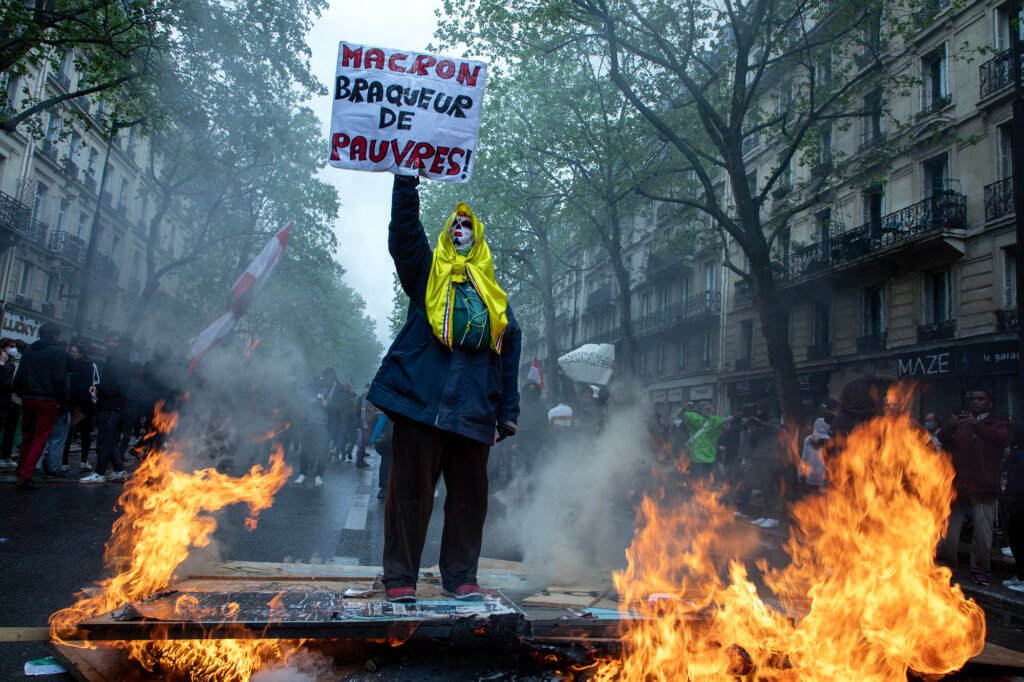 During the May Day demonstration in Paris, a protester stands surrounded by flames, holding a sign that reads, 'Macron, robber of the poor.'