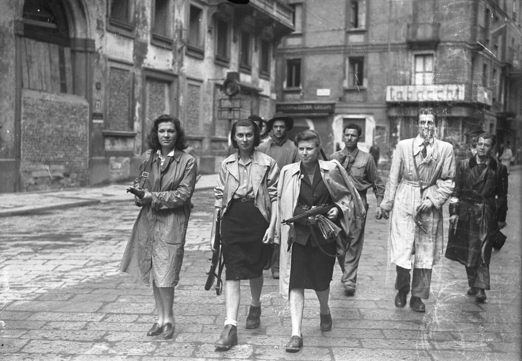 Three partisan women returning guns to the Allies after the Liberation, with militants of GAP (Gruppi di Azione Patriotica) on their background.