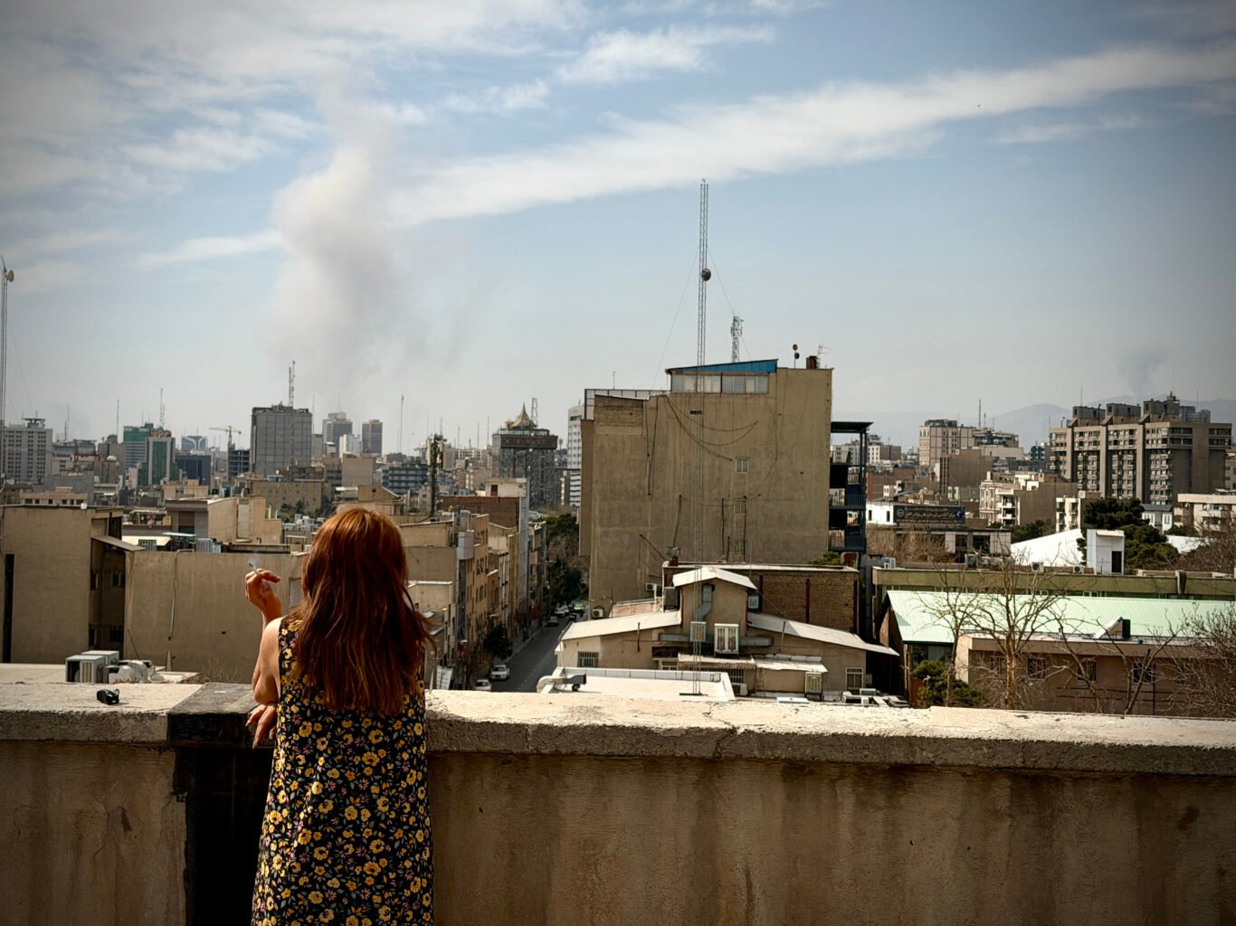 A woman smokes a cigarette on a rooftop as explosions from joint US-Israeli strike show on the background in Tehran.